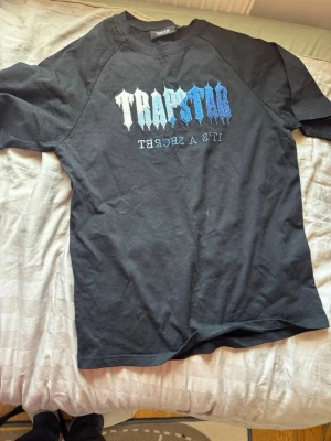 Trapstar svart t-shirt med blått tryck - Svart t-shirt från Trapstar med stort tryck i vitt och blått på bröstet där det står 'TRAPSTAR' och 'IT'S A SECRET' spegelvänt. Klassisk rund hals och korta ärmar. Perfekt för dig som gillar streetwear och vill sticka ut.