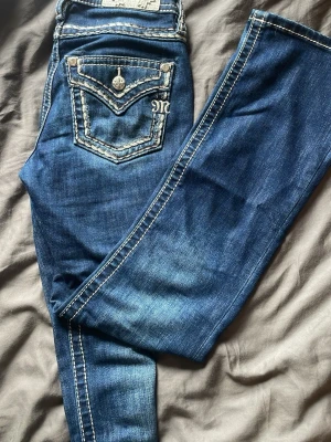 Mörkblå Miss Me jeans med kontrastsömmar - Säljer ett par mörkblå Miss Me jeans med tydliga kontrastsömmar och snygga detaljer på bakfickorna, inklusive metallknappar och broderad logga. Jeansen har en klassisk femficksmodell och är tillverkade i stretchigt denimtyg för skön passform.
