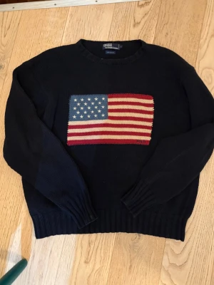 Mörkblå stickad tröja Polo Ralph Lauren - Säljer en mörkblå stickad tröja från Polo Ralph Lauren med en stor amerikansk flagga broderad på bröstet. Tröjan har rund halsringning och ribbade muddar vid ärmslut och nederkant. Klassisk och tidlös design i mjukt material.
