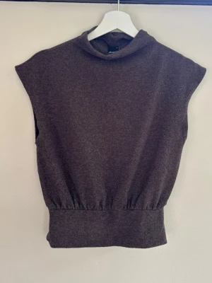 Brun ärmlös stickad topp Gina Tricot - Mysig brun ärmlös stickad topp från Gina Tricot med hög krage och bred ribbad kant nedtill. Perfekt att styla över en skjorta eller t-shirt för en trendig lager-på-lager-look. Materialet känns mjukt och stretchigt.