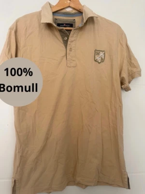 Beige kortärmad piké - Klassisk beige pikétröja i 100% bomull. Tröjan har korta ärmar, krage och knappar framtill samt en broderad patch på bröstet. Perfekt till en avslappnad stil.