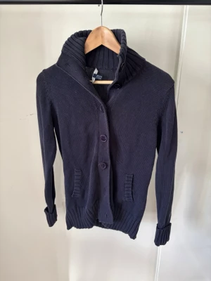 Gant Cardigan - Mörkblå stickad kofta med stora knappar framtill och bred ribbad krage. Koftan har långa ärmar, ribbade muddar och två fickor framtill. Perfekt för lager-på-lager och en chill stil.