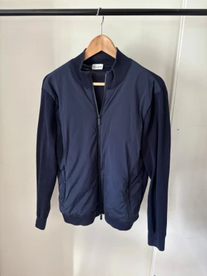 John Henric - Cardigan - Snygg mörkblå Cardigan med dragkedja framtill och ribbade muddar vid krage, ärmslut och nederkant. Jackan har släta ärmar och sidofickor. Materialet ser ut att vara en mix av polyester och bomull, vilket ger en sportig och clean look.
