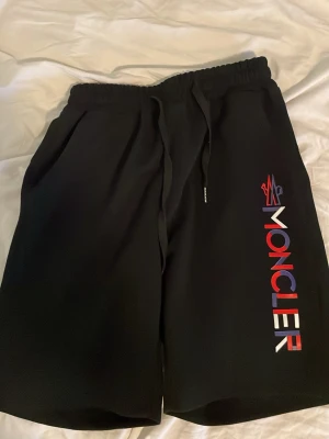 Svarta Moncler shorts med tryck - Svarta shorts från Moncler med färgglatt logotyptryck längs ena benet. Modellen har elastisk midja med snörning och sidofickor. Perfekta för en avslappnad och trendig stil.