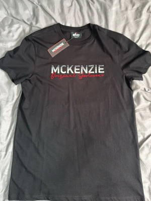 Svart McKenzie t-shirt med tryck - Svart t-shirt från McKenzie med stort tryck på bröstet i vitt och rött där det står 'McKenzie Original Sportswear'. Klassisk rund hals och korta ärmar. Perfekt för en avslappnad och sportig stil.