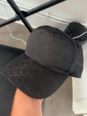 Svart keps med diskret mönster - Säljer en svart keps med böjd skärm och diskret ton-i-ton mönster över hela kepsen. Kepsen har en klassisk form och justerbar rem baktill. Perfekt för dig som gillar stilrena accessoarer med subtila detaljer.