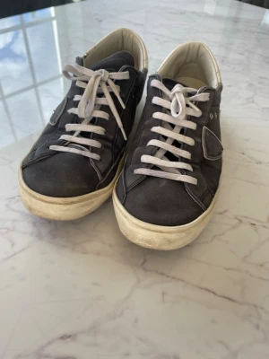 Philippe Model grå låga sneakers - Grå sneakers från Philippe Model med vit sula och vita skosnören. Skorna har en ikonisk sköldlogga på sidan och flera metallöglor för ventilation. Ovandelen är i mocka och insidan är ljus. Perfekta för en avslappnad och trendig look.