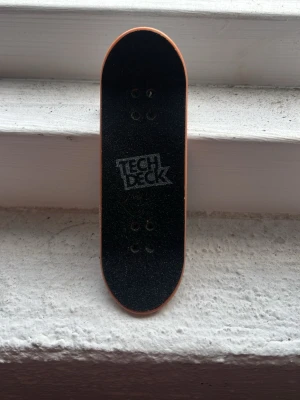 Tech Deck fingerboard svart - Säljer en Tech Deck fingerboard med svart grepptejp och logga på ovansidan. Brädan har en klassisk skateboardform med vita hjul och träfärgad kant. Perfekt för dig som gillar att göra tricks med fingrarna!