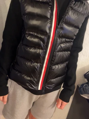 Moncler cardigan - Snygg svart dunjacka från Moncler med glansig front och stickade svarta ärmar. Jackan har två dragkedjeförsedda fickor och en röd-vit rand längs dragkedjan. Säljer då den ej passar mig, Scanningen på själva moncler märket funkar ej. Snabb affär