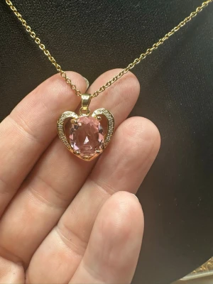 Halsband med hjärthänge och rosa sten - Guldigt halsband med ett hjärtformat hänge som har en stor rosa sten i mitten. Hänget är dekorerat med små gnistrande stenar runt hjärtat för extra bling. Kedjan är tunn och elegant, perfekt för att ge en romantisk touch till din outfit.