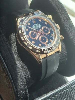 Role klocka - En klocka med svart urtavla, guldiga detaljer och svarta subdials. Boetten är i guld och armbandet är svart och i syntetmaterial. Klockan har kronograf och tydliga indexmarkeringar i guld. Perfekt för dig som vill sticka ut med en lyxig accessoar. Klockan har automatiskt urverk.