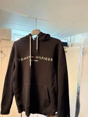 Svart hoodie från Tommy Hilfiger - Snygg svart hoodie från Tommy Hilfiger med vit logga och text på bröstet. Klassisk huva med vita dragsnören och stor magficka framtill. Perfekt för en avslappnad och trendig stil.