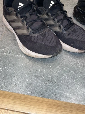 Svarta Adidas sneakers med mesh - Säljer ett par svarta Adidas sneakers med klassiska tre ränder på sidan. Ovandelen är i meshmaterial för bra andningsförmåga och sulan är vit med lätt struktur. Skorna har svarta snören och låg profil som passar till många stilar.