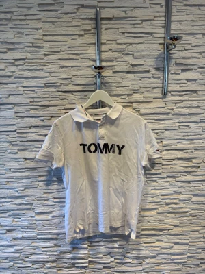 Vit pikétröja med svart TOMMY-tryck - Vit pikétröja med klassisk krage och korta ärmar. På bröstet finns ett stort svart tryck med texten 'TOMMY'. Tröjan har en enkel och stilren design, perfekt för en avslappnad look.