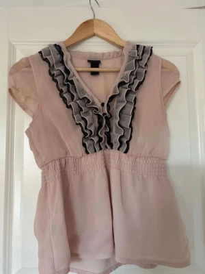 Ljusrosa blus med volang från H&M - Supersöt ljusrosa blus från H&M med korta ärmar och volangdetaljer i svart och grått längs v-ringningen. Blusen har resår i midjan för en snygg passform och är gjord i ett tunt, luftigt material som känns lätt på kroppen.