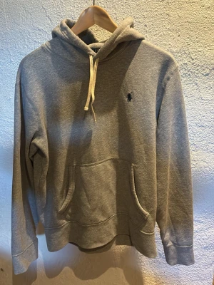 Grå hoodie med broderad logga - Klassisk grå hoodie med huva och vita dragsnören. Har en känguruficka framtill och en liten broderad logga på bröstet. Tillverkad i mjukt sweatshirtmaterial, perfekt för en avslappnad och stilren look. Hör av er vid frågor😄
