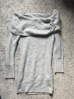 Grå offshoulder stickad klänning - Snygg grå stickad klänning med off shoulder-design och bred krage. Tröjan har dekorativa snörningsdetaljer på ena sidan av kragen och långa ärmar med ribbade muddar. Perfekt för dig som gillar trendiga och bekväma plagg.