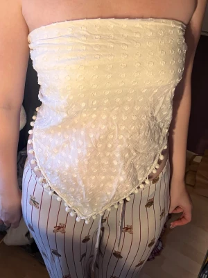 Vit axelbandslös bandeau topp - Säljer en vit axelbandslös bandeau topp med strukturmönster och små bollar längs kanten. Toppen knyts i ryggen och har en triangelformad nederdel. Perfekt för sommarens varma dagar och ger en trendig look.