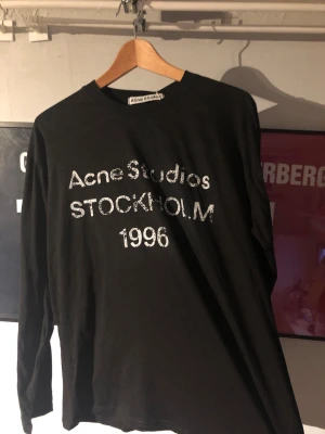 Svart långärmad topp Acne Studios - Cool svart långärmad topp från Acne Studios med vit text 'Acne Studios STOCKHOLM 1996' framtill. Klassisk rund halsringning och relaxed fit. Perfekt för dig som gillar streetwear och enkel stil.