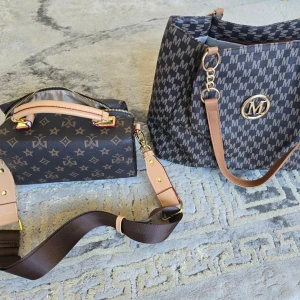 Brun handväska med M-mönster och guldiga detaljer - Snygg brun handväska med upprepade M-mönster och stora guldiga detaljer. Väskan har beige axelremmar i konstläder och en rymlig insida med flera fack. Perfekt för dig som vill ha plats för allt du behöver. Stängs med dragkedja och har en modern bucket-form.