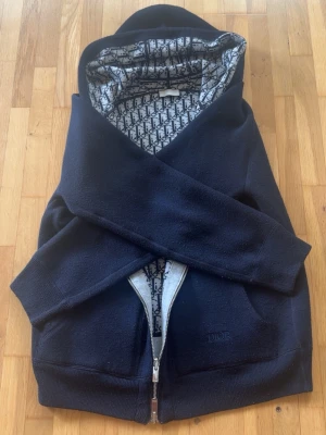 Marinblå D I O R hoodie med monogramfoder - Säljer en marinblå hoodie från D I O R med dragkedja och två stora fickor framtill. Insidan är fodrad med D I O R S ikoniska monogrammönster i grått och svart. Huvan har dragsko med metalländar och materialet känns mjukt och lyxigt. Perfekt för dig som gillar exklusiva streetwear-plagg.