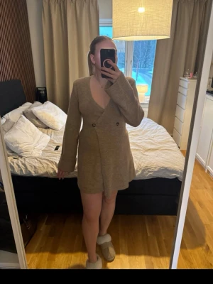 Beige omlottklänning, Adoore - Kort beige omlottklänning med v-ringning och långa ärmar. Klänningen har en figurnära passform och stängs med två knappar framtill. Perfekt för dig som gillar en enkel men stilren look.