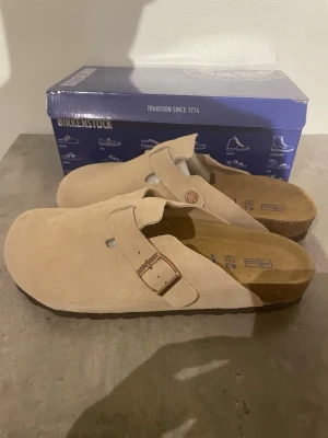Beige mockatofflor Birkenstock - Säljer ett par klassiska beige tofflor från Birkenstock i mjuk mocka. Modellen har täckt tå, justerbart spänne i metall och korksula för extra komfort. Perfekta för dig som vill ha både stil och bekvämlighet.