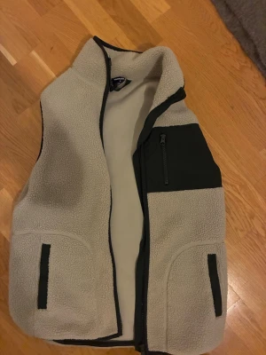 Beige fleeceväst från Newbody XS - Mysig beige fleeceväst från Newbody i storlek XS. Västen har svarta detaljer vid fickor och bröstficka med dragkedja. Tillverkad i 100% polyester och perfekt för lager-på-lager-stil. Hög krage och hel dragkedja framtill.