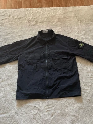 Stone Island jacka  - Snygg svart overshirt från Stone Island med dragkedja framtill och två stora bröstfickor med lock. Klassisk krage och ikonisk patch på vänster ärm. Tillverkad i ett lätt, strukturerat material som ger en modern look. Vi kan diskutera priset.