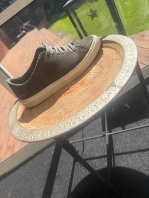 Arigato Mörkgråa låga sneakers i mocka - Snygga mörkgrå sneakers i mocka med klassisk låg profil och vita sulor. Skorna har matchande mörkgrå skosnören och en stilren design utan synliga loggor. Perfekta för dig som gillar en clean och enkel look.