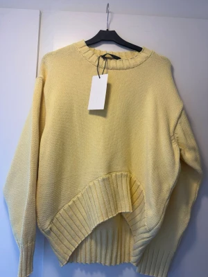 Gul stickad tröja från ZARA - Trendig gul stickad tröja från ZARA med oversized passform och asymmetrisk ribbad nederkant. Tröjan har rund halsringning och långa ärmar. Perfekt för dig som vill ha en unik och bekväm look. Materialet känns mjukt och skönt mot huden.