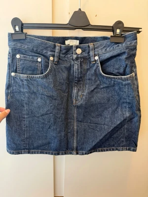 Mörkblå jeanskjol från Arket i klassisk modell - Snygg mörkblå jeanskjol med fem fickor och klassisk knappstängning framtill. Kjolen har en rak passform och är tillverkad i denim med tydliga sömmar. Perfekt att styla med en t-shirt eller hoodie för en avslappnad look.