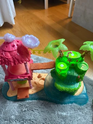 Hatchimals lekset tropisk ö - Färgglatt lekset från Hatchimals med tropiskt ö-tema. Innehåller en rosa hydda, gröna palmer och bro, samt detaljer som vatten och sand. Perfekt för fantasifull lek och samling av småfigurer.