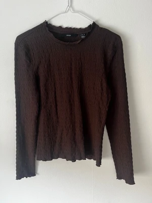 Brun långärmad topp från Vero Moda - Brun långärmad topp från Vero Moda med ribbad struktur och vågig kant vid halsringningen. Toppen har en tight passform och är tillverkad i ett stretchigt syntetmaterial 