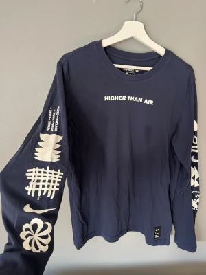 Blå långärmad Nike Dri-Fit T-shirt - Snygg långärmad blå Nike Dri-Fit T-shirt med trycket 'HIGHER THAN AIR' på bröstet och coola vita grafiska tryck längs ärmarna. Perfekt för dig som gillar streetwear och sportig stil.