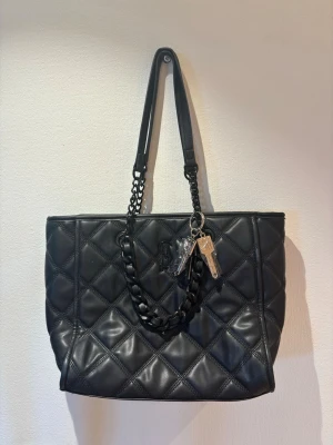 Svart quiltad handväska från Steve Madden - Snygg svart handväska från Steve Madden i quiltad design med kedjedetaljer på axelremmarna. Väskan har ett diskret SM-monogram framtill och nyckelringar med metallnycklar som accessoar. Tillverkad i konstläder med en rymlig form.