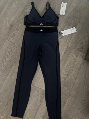 Alo sportiga bodys set i svart - Säljer två snygga bodys set från Alo, båda i svart med kontrasterande sömmar. Seten består av sport-bh och höga tights med logga i midjan. Materialet är stretchigt och följsamt, perfekt för en aktiv livsstil. Stilsäkra detaljer med vita och svarta linjer för en modern look.