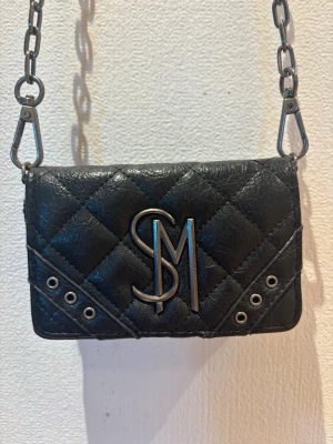 Svart quiltad axelväska från Steve Madden - Snygg svart axelväska från Steve Madden med quiltat mönster, silverfärgade detaljer och kedjerem. Väskan har stora SM-loggan framtill och dekorativa nitar. Perfekt för dig som gillar en edgy och trendig look.