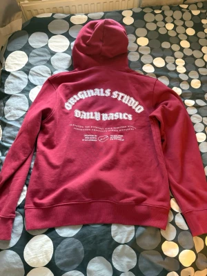 Vinröd hoodie Originals Studio - Snygg vinröd hoodie från Originals Studio med vit text på bröstet och ryggen. Klassisk känguruficka framtill och justerbar huva. Tillverkad i mjukt material, perfekt för chill dagar. Texten på ryggen ger en cool streetwear vibe.