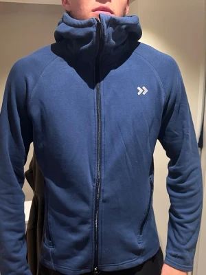 Blå hoodie med dragkedja - Mörkblå hoodie med hel dragkedja framtill och hög krage. Tröjan har två sidofickor med dragkedja och en liten vit logga på bröstet. Tillverkad i mjukt material och har en huva som kan dras upp för extra värme. Perfekt för chill eller träning.