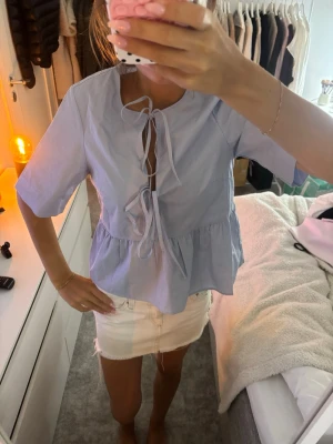 Ljusblå blus med knyt och volang - Supersöt ljusblå blus med korta ärmar, knytning framtill och volang nedtill. 