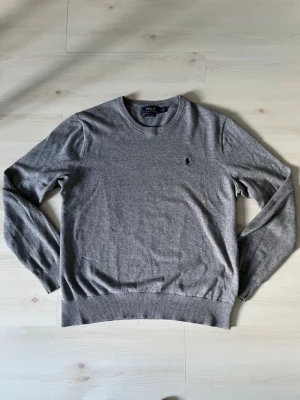 Grå stickad tröja Polo Ralph Lauren - Snygg grå stickad tröja från Polo Ralph Lauren i mjuk pima bomull. Tröjan har rund halsringning, långa ärmar och den klassiska broderade loggan på bröstet. Perfekt till jeans eller chinos för en clean look.