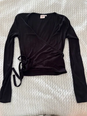 Svart omlottopp med långa ärmar - Snygg svart omlottopp med långa ärmar och knytband i sidan. Toppen har en djup v-ringning och är tillverkad i ett mjukt, stretchigt material som sitter skönt på kroppen. Perfekt att styla med jeans eller kjol för en trendig look.