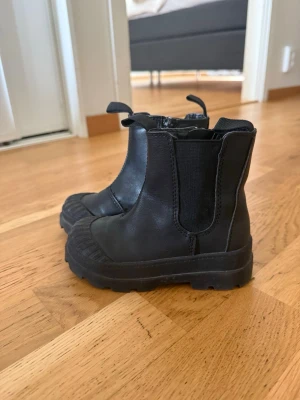 Svarta Chelsea boots med grov sula - Säljer ett par svarta Chelsea boots i skinnimitation med elastiska paneler på sidorna och dragkedja på insidan. De har en chunky, räfflad sula och dragflikar både fram och bak. Perfekta för dig som gillar en tuffare stil och vill ha bekväma boots till hösten.