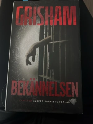 Bekännelsen av John Grisham - Spännande thrillerroman av John Grisham med svart omslag och röd text. Boken handlar om en präst och en före detta fånge som försöker avslöja sanningen bakom ett brutalt brott i Texas. Perfekt för dig som gillar nervkittlande berättelser och rättsdrama.