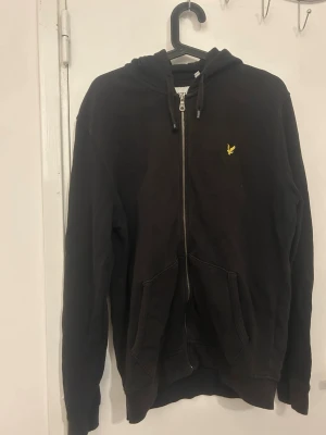 Svart zip-hoodie med loggan lyle & scott - Säljer en svart hoodie med dragkedja och huva. På bröstet finns en liten gul broderad logga. Hoodien har två fickor framtill och ribbade muddar vid ärmar och nederkant. Perfekt för en chill och avslappnad stil.