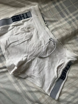 Vita shorts från Tommy Hilfiger - Säljer ett par vita shorts från Tommy Hilfiger med marinblå detaljer och bred resår i midjan med snörning. Shortsen har fickor på sidorna och ett sportigt band längs benen. Perfekta för sommaren!