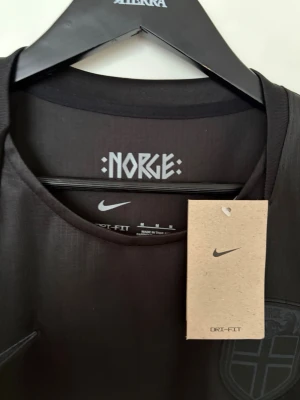 Svart Norge fotbollströja Nike - Säljer en helt ny, snygg svart fotbollströja med Norge-tryck och emblem på bröstet. Tröjan är tillverkad i Dri-FIT-material som andas och har rund halsringning. Perfekt för träning eller match. Finns både S och M i storlek 