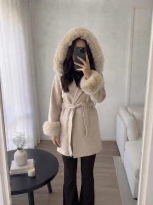 Beige kappa med pälsdetaljer  - Säljer en elegant beige kappa med äkta päls på huva och ärmslut. Kappan har en knytning i midjan och är perfekt för kyligare dagar. Den har en mjuk och lyxig känsla, vilket gör den till ett stilrent val för höst och vinter. S/M