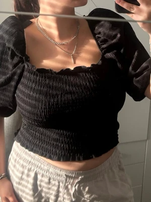 Svart croppad blus med puffärm - Trendig svart croppad blus med smockad struktur och puffiga korta ärmar. Fyrkantig urringning ger en snygg siluett och toppen har en figurnära passform. Perfekt att styla med både jeans och kjol för en cool look.